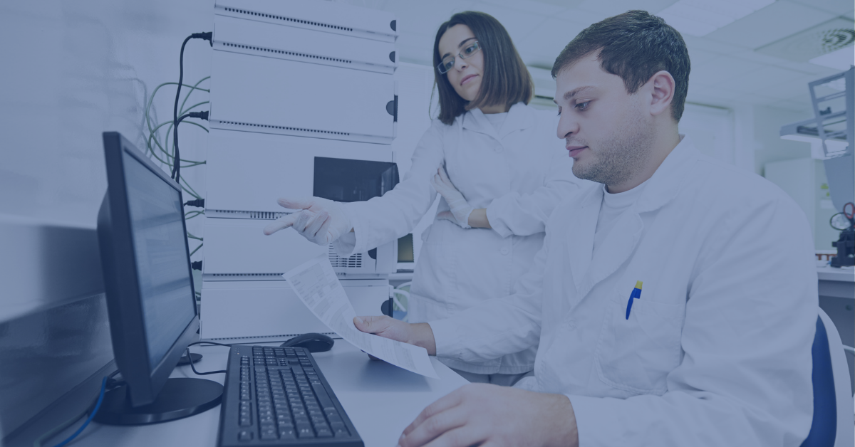 Two scientists in lab coats reviewing validation documents on a computer screen, with laboratory equipment in the background.