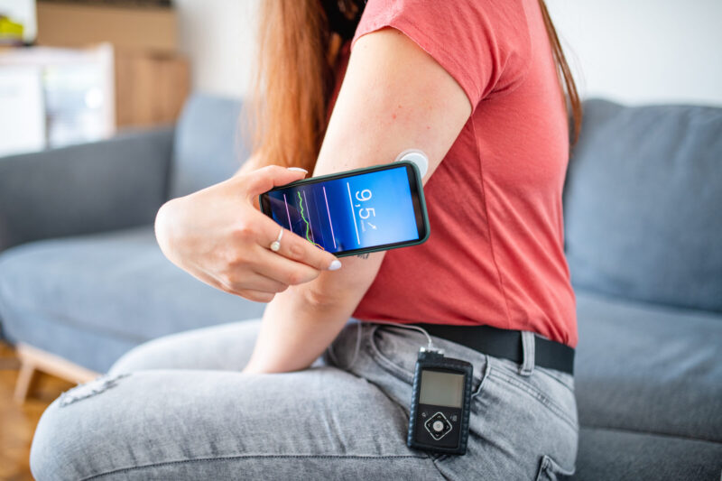 Side view of Caucasian woman wearing casual clothing and insulin pump at her waist sitting on sofa and swiping sensor to determine glucose measurement.