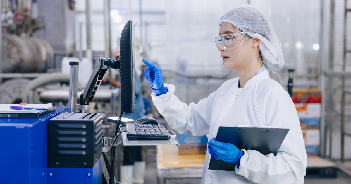 A person in a white lab coat, hair net, and blue gloves inspects a monitor and control panel at a manufacturing workstation in a bright industrial facility.